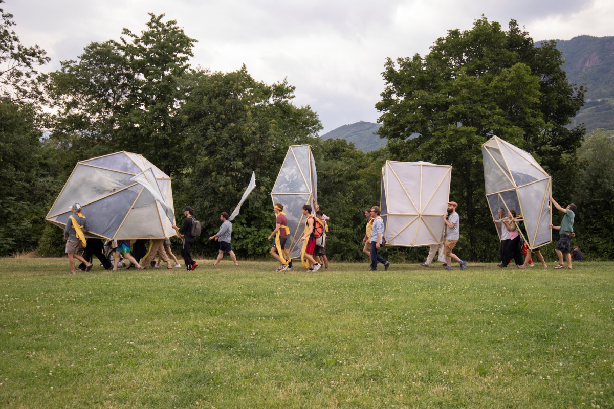 Eine Gruppe von Menschen tragen mehrere überdimensionale Schirme über eine Wiese