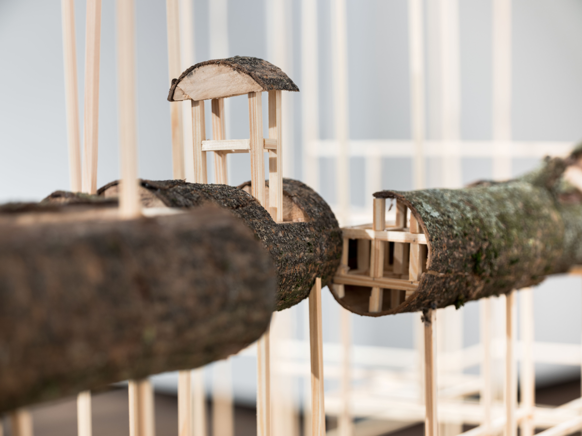 Installation aus dicken Ästen, die wie ein Baumhaus gestaltet ist. Im Querschnitt sind Holzstäbe in einem schachbrettartigen Muster übereinander angeordnet. Das Ganze wird von weiteren Holzstäben gestützt.