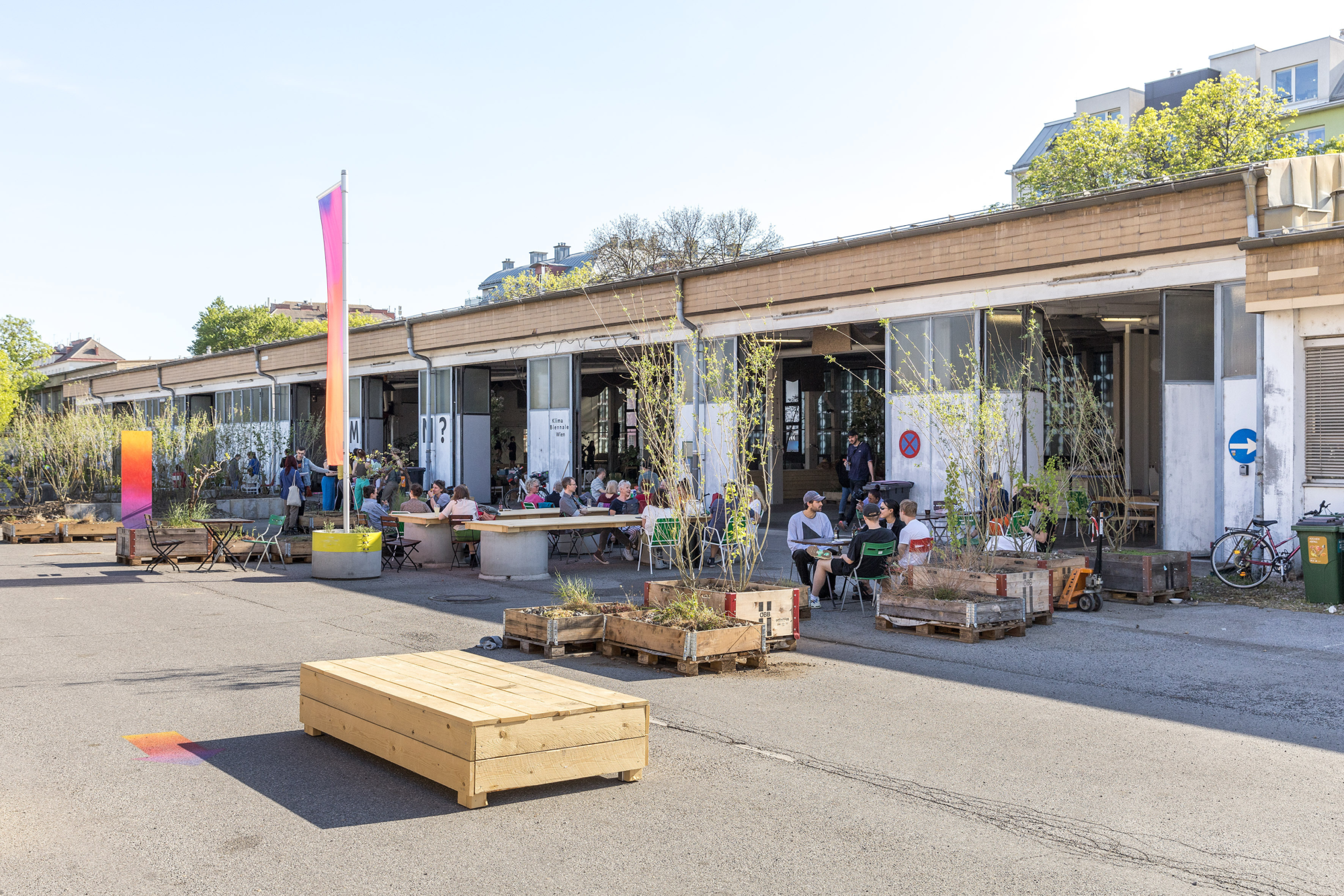 Asphaltierter Platz mit Halle, davor Pflanzen und Sitzmöglichkeiten