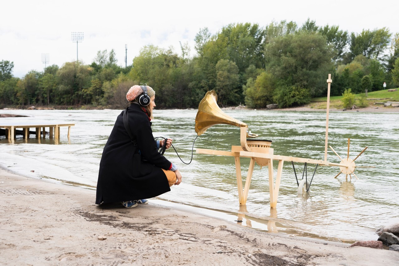 Ein Person sitzt vor einen umgebauten Gramophon vor einem Fluss und belauscht das Wasser