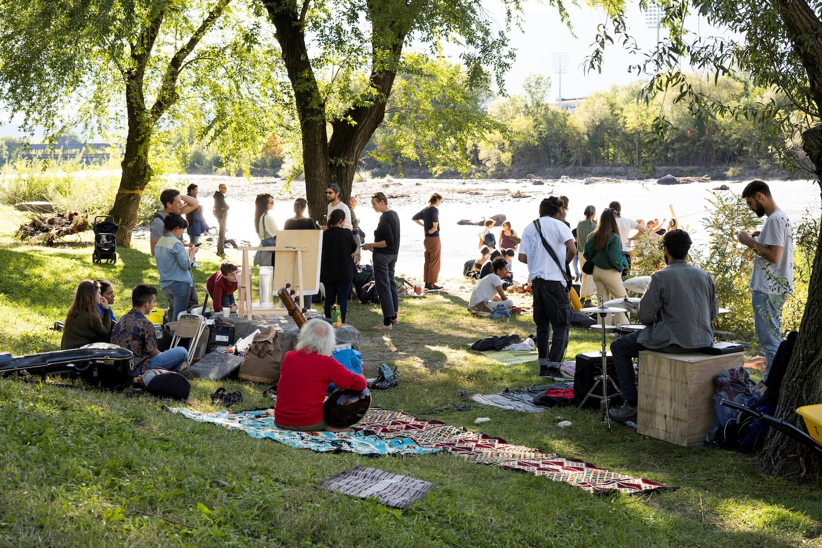 Eine Gruppe an Menschen sitzt im Schatten von Bäumen und schaut auf einen Fluß