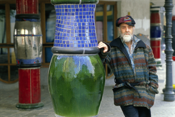 Mann mit weißem Vollbart und Kappe und bunter Jacke lehnt an einer grün-blauen Säule